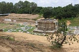 A ruined ancient Indian temple set in a shallow valley of sand, surrounded by trees at a distance.