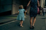 A girl, perhaps five years old, with blonde, curly hair and wearing a blue dress, is walking down a street. She is holding the hand of a woman in a blue dress.