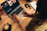 Woman sitting at a desk editing a video on a laptop. There is also a camera and coffee on the desk.