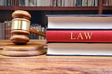 Wooden judge’s gavel resting on its sound block beside a stack of hardcover law books — one with a prominent red spine labeled “LAW” — on a polished wooden desk, framed by a blurred background of law-library shelves.