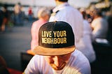 A close up photograph of a man wearing a black and yellow cap with the words ‘love your neighbour’ written over the brim