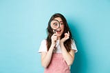 Happy young woman looking through a large magnifying glass with an excited face as if she has just discovered something. She is standing up, wearing pink dungarees and a white T-shirt against a blue background.