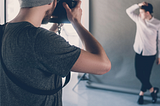 A photographer in a studio captures a male model posing against a gray backdrop during a professional photoshoot.