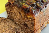 upside down banana bread with a slice cut, on a white plate sitting on a textured bright yellow background