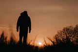 Rear view of a man walking through a field, toward the setting sun.