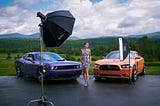 Behind the scenes of a fashion photoshoot showing flash equipment, model, and lots of detail in a Vermont summer scene with two modern muscle cars.