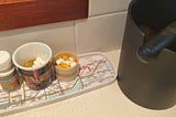 Tray on a kitchen bench with pills in small cups, next to a coffee grind waste container