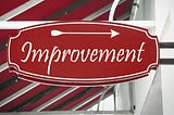 Red store sign that says “Improvement” in script with an arrow pointing to the right