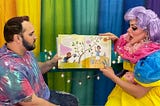 Two Drag Queen Story Time presenters are holding up a children’s book in front of a rainbow background as they read the story aloud