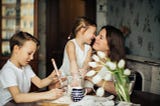 A mother with dark brown hair smiles as she hugs her brown-haired daughter. Her brown-haired son sits beside them at a table. There are white flowers on the table.