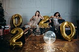 What to do when your New Year’s resolutions fail. Image: Women on couch drinking wine surrounded by large year 2020 balloons.
