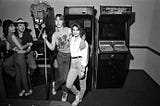 A young couple stand with their arms around one another in front of a pair of upright video games, while another couple lean against a pinball machine