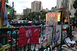 Image is a photograph of a subway station entrance/exit in downtown Manhattan by City Hall covered in hand painted signs about police brutality and abolishing the police. Text on signs includes: “Oink! Oink! Oink! Cops are Killers”, “Abolish the Police”, “Black Lives Matter”, and “Justice for Layleen Polanco”. Further in the distance beyond the subway entrance are a crowd of people gathered. Beyond the people are high rise buildings.