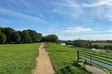 A gravel footpath stretching ahead between two grassy banks. It runs alongside the River Parrett, which is to the right of the picture. There are riverside benches and trees in the distance. It’s a sunny day with blue sky and wispy white clouds. This was taken at the very end of my half marathon run.
