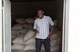 A African man looks down at the smartphone in his hand at the entrance of a storefront full of bags of grain
