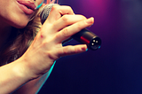 Singer sings into microphone held by their hand. Only their mouth is visible. Dark background with purple hue.