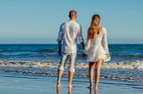 An attractive young couple, both dressed in white, facing a stunning blue ocean