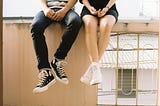 A man and woman sitting on a wall. Only their legs are visible, with both wearing Converse sneakers.