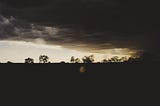 A silhouette of trees in the horizon with dark storm clouds rolling in from the right.