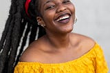 Smiling black woman wearing a bright yellow blouse. She is also wearing dreadlocks.