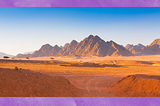 A panoramic view of a jagged mountain range from the point of view of a mile away in an orange-dirt desert.