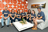 Seven members of the Architectural Designs team pose in an office with a bright orange and write wall. They are wearing blue shirts that read Bueno to honor AD alumnus Ryan Bard, who is pursuing his lifelong dream of becoming a commercial pilot.