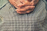 an older white woman’s hands folded in her lap, on the background of a black and white checked skirt.