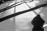 Black and white photo of shadows of trees criss-crossing a field of snow. The shadow of a human figure is in the lower right-hand corner of the picture.