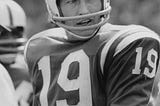 A black-and-white photo of Johnny Unitas in a Baltimore Colts uniform with the number 19 on his uniform. He is wearing a football helmet with the Baltimore Colts logo partially visible. His left arm is bent in front of him.