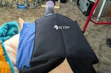 A person is lying down with their leg elevated and wrapped in a black therapy device, indicated by the label “SM FOLD THERAPY.” Surrounding them are various household items, including a blue blanket and a red walker.