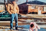My father, a young brown-skinned man with black hair and glasses, is looking down and smiling at me as a toddler bending down to touch the bits of snow left on the ground. He is wearing a brown jacket and blue jeans and has his hands in his pockets. I am wearing a pink puffy snowsuit. We are in the front yard of a suburban neighborhood with brick ranch homes.
