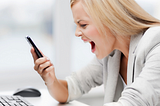 Frustrated blonde woman in a grey suit jacket yelling at her cell phone.