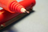 Picture shows a close up of a red pen with the cap removed. The pen is resting on the cap such that the point is raised.