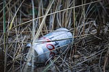 A discarded silver can of Coors Light beer lies on the ground amidst weeds and dirt.