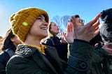 a person in a yellow knit hat, facing right, smiling and clapping, amid a crowd of people