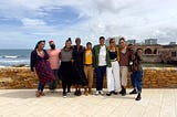 A group of Black trans and gender-expansive folks smiling and standing on payment close to a shore with a low dark yellow brick wall.