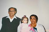 A baby, middle, wears a pink outfit and sits in the arms of her grandmother, a dark-skinned woman with a black bun and colorful saree. A man, the baby’s grandfather, stands to the left, wearing a white collared shirt and black vest.