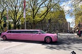 A bright pink limousine about to go across an intersection on an urban street with trees in the background on a sunny day.