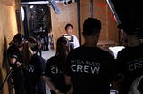 Actor Tony Leung Chiu-wai is in a warehouse. A camera crew of 5 wearing black t-shirts with media studio logo is shooting the interview.