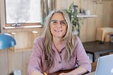 Photo of the author, Uma Bode, sitting at her desk facing the camera, smiling, holding a pencil in her hand. The background is out of focus and shows a window, a plant, and shelves.