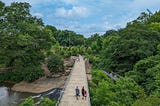 Pedestrians walking over a bridge crossing a river and bordered by green trees