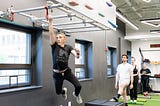 A man swings by one arm on monkey bars, wearing a black Amplify Fitness shirt and looking focused at the bar in front of him. Three people wait in the background for their turn.