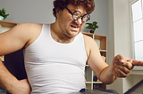 Man sitting in a white sleeveless undershirt and pj’s looks and points angrily at his computer.