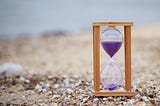 Close-up photo of a wooden hourglass with purple sand in it. Background is of a beach.