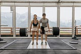 Image shows young woman and man in athletic wear lifting a large barbell together in a gym. The wall behind them is full of windows with view of mountains.