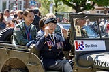 An old man with war medals waves from a jeep with a young citizen seated behind