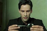 Keanu Reeves as Neo, wearing a black suit and shirt, looks at a spoon filmed in such a way that at a glance it looks like a key.