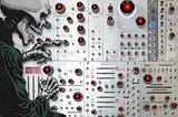 A complex control panel whose knobs have all been replaced with the menacing red eye of HAL9000 from Kubrick’s ‘2001: A Space Odyssey.’ A skeletal figure on one side of the image reaches out a bony finger to twiddle one of the knobs. Image: Cryteria (modified) https://commons.wikimedia.org/wiki/File:HAL9000.svg CC BY 3.0 https://creativecommons.org/licenses/by/3.0/deed.en — djhughman https://commons.wikimedia.org/wiki/File:Modular_synthesizer_-_%22Control_Voltage%22_electronic_music_shop_in