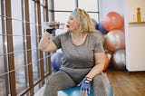 Larger neckered looking woman sitting on an exercise ball at the gym drinking from a water bottle.