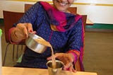 A woman pours filter coffee from a rimmed bowl to a rimmed glass. She is smiling. A bowl of something savory and yummy sits in front of her.